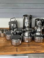 Photo of 10 stainless steel houseware items arranged on a wooden surface including pots, canisters and a multi-tiered container