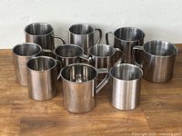 11 stainless steel mugs in various sizes with handles, arranged on a wooden surface against a white wall.