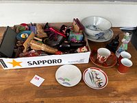 Photo showing the entire lot with many decorative ceramic items, figurines, bowls, boxes, and trinkets spread out on a table and contained inside an open box labeled 'SAPPORO'.
