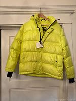 Full front view of bright yellow TNA puffer jacket showing hood, pockets, and front closure.