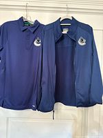 Photo of two navy blue Vancouver Canucks shirts hanging on hangers on a door. Both shirts display the team logo on the chest area.