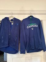 Two Vancouver Canucks hoodies hanging on hangers side by side; a zip-up and a pullover.