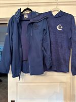 Two garments, a jacket and a pullover, hanging on hangers showing front with Vancouver Canucks logos.