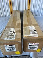 Photo showing two unopened brown cardboard boxes, each containing an AutoShack Air Suspension Strut Model KAS229M29F. Boxes positioned side by side on table.