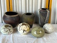 Front view of seven handmade decorative ceramic and pottery vases and vessels arranged on a white cloth with curtain background. Three taller textured earth toned vases at back with four orb-like decorated vessels in front.