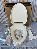 Full view of bidet toilet seat with lid and seat open, hoses, and packing materials around.