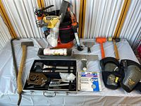 Wide view of all tools laid out on a white cloth, showing axe, crowbar, caulking gun, putty knife, mallet, trowel, knee pads, metal trays with smaller tools, and painting related items.