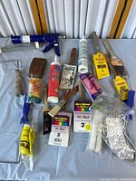 Overview of painting and caulking tools laid out on a table showing caulking gun, multiple paint brushes, paint roller with covers, and putty knife.