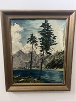 Framed oil painting of pine trees on a small island by a lake with gray mountains and partly cloudy sky in the background. Signed lower left. Frame is bronze/gold tone with some wear.