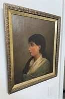 Oblique view of framed oil painting depicting profile portrait of a young woman, gold wood frame.