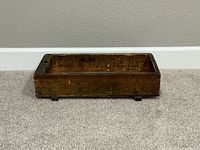 Rectangular antique/vintage wooden box with visible wear and stamped text on front, sitting on carpeted floor.
