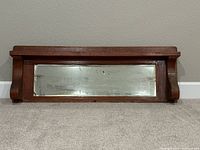 Front view of antique wooden mirror with shelf resting on floor showing full view of mirror and corbels.