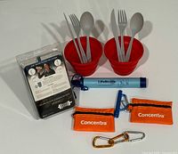 Photo of two collapsible red bowls each containing a set of grey utensils (fork, spoon, chopsticks), Sawyer water filtration system packaging, LifeStraw, two orange small first aid packs labeled Concentra, whistle and carabiners.