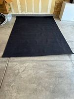 Top-down photo showing the full black rug on concrete floor, edges mostly intact with one corner slightly bent.