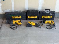 Three DeWalt power tools and three DeWalt carrying cases lined up on concrete floor.