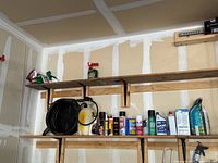Two wooden shelves mounted on a garage wall with various items including spray cans, bottles, bags, and boxes of automotive and garage-related products.