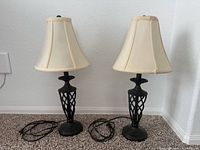 Two black iron table lamps with beige shades placed side-by-side on carpet against white wall.