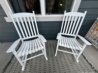 Two white wood rocking chairs side-by-side on front porch carpet with visible weathering on armrests and seats.