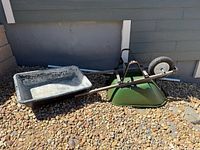 Two wheelbarrows including one with a plastic mixing tub behind them on gravel next to a building.