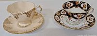 Two antique Royal Albert tea cups and saucers displayed side by side on a white surface; one set white with gold patterns, the other with black and floral gold designs.