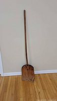 Full view of the tall wooden paddle against a wall, showing the entire length including the handle and blade, resting on a hardwood floor.