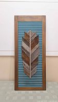 Full view of wooden feather wall art with teal blue slatted background and brown wood feather in center, framed in medium brown wood.