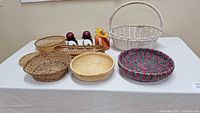Eight woven baskets displayed on a white table with a small fabric pumpkin decoration. Variety includes natural, white, and multicolored patterns, different shapes and weaving styles.