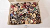Photo showing a large quantity of assorted buttons in a white box, various colors, sizes, and materials