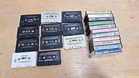 Top view of 15 vintage cassette tapes in plastic cases and loose cassettes on a wooden surface.