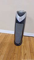 Full view of GermGuardian tower air purifier standing on wooden floor against a wall, showing front and base detail.