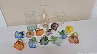 Overview of 10 colorful blown glass flower napkin holders with green stems and 4 glass pitchers behind them on a white surface.
