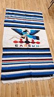 Full view of rectangular hand woven Mexican blanket laid flat on wooden floor, showing central eagle motif and 'CANCUN' lettering with striped pattern.