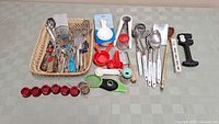 Wide view of assorted kitchen gadgets and tools including red measuring cups, metal utensils, and peelers in a basket and spread out