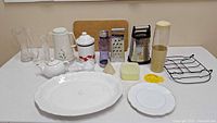 Full lot of kitchen items arranged on a table including platters, vases, cutting board, graters, canisters, teapot, soap, and utensils.