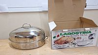 Stainless steel roaster with dome lid partially lifted, shown with original box.