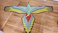 Top view of large bird-shaped kite laid flat on wooden floor showing full wingspan and colorful feather pattern.