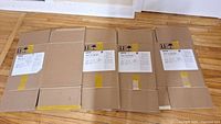 Four flat-packed medium size cardboard boxes with Mayzo brand packaging labels and yellow tape reinforcement laid out on wooden floor.