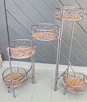 Full view of multi-tier metal and wicker/rattan plant stand showing four tiers and five plant holders, gray metal with natural wicker/rattan woven bases.