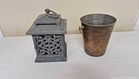 Photo of a heavy metal candle holder with intricate openwork and a metal bucket with side handles on a white surface