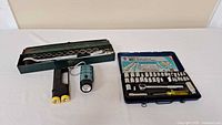 Two ratchet socket sets and three portable flashlights displayed on a white surface. One socket set in a green metal case with sockets visible, the other in a black plastic case with sockets and tools visible.