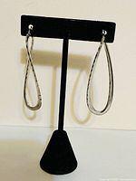 Front view of the pair of teardrop-shaped sterling silver earrings displayed on a black stand, showing their size and mobius twisted shape with notched edges.