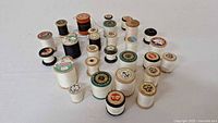 Photo showing a collection of 24 vintage wooden spools of thread in white and black, with various brands visible labeled on top.