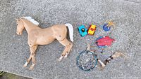 Photo showing beige plastic horse with articulated legs, white mane and tail, two small toy vehicles, a red crab, a blue figurine, and a dreamcatcher with feathers and beads on concrete surface.
