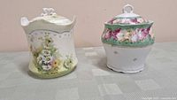 Photo shows two ceramic biscuit barrels placed side by side on a tablecloth. The left barrel is decorated with daisies and other small flowers and has a scalloped top with a handle shaped like a flower. The right barrel features a green band near the top and a floral pattern of pink roses.
