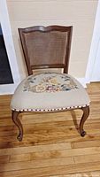 Front view of the vintage wooden chair showing the cane backrest and needlepoint floral seat upholstery, resting on wooden flooring.