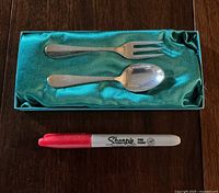 Two child-sized sterling silver cutlery pieces, fork and spoon, displayed on green satin in a box, with a red Sharpie marker for scale.