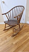 View of the Windsor style rocking chair showing curved spindled back, solid contoured wood seat, turned legs, and rockers on hardwood floor.