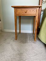 Single oak nightstand with one drawer and round wooden knob, standing on carpeted floor next to bed.