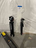 Two black tripods standing upright against a plastic covered wall with a blue taped paper above them. Box for the Solidex tripod on the floor shows it is included in the lot.