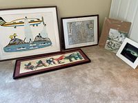 Five assorted pieces of art arranged on floor; includes three framed pieces (stylized airplane scene, map, native-inspired 'EARTH' art) and two unframed artworks (nature scene and blue-toned photo/print).